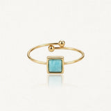 Bague ouverte dorée avec une pierre carrée bleu-vert au centre et des billes aux extrémités.