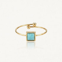 Bague ouverte dorée avec une pierre carrée bleu-vert au centre et des billes aux extrémités.