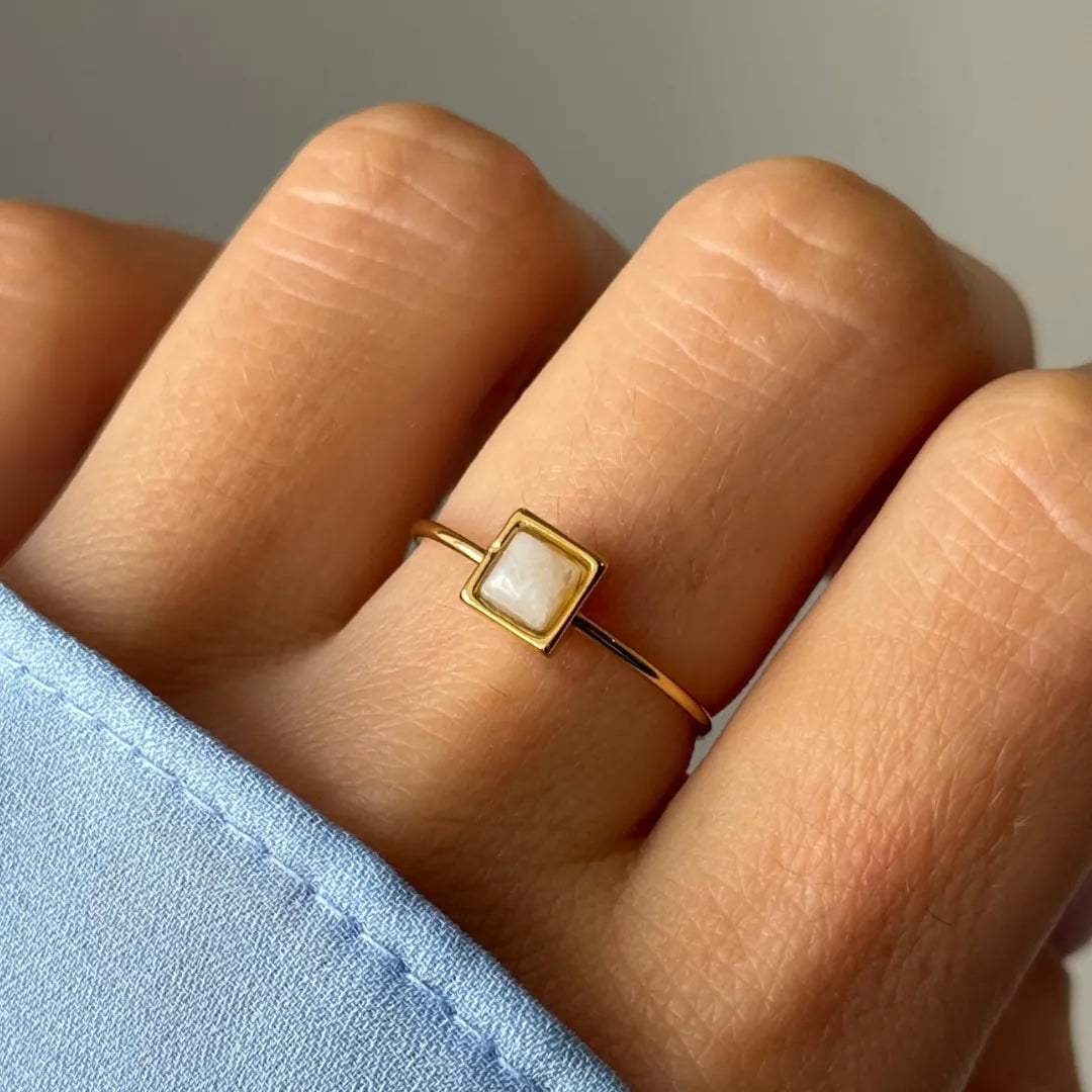 Bague fine dorée avec un motif en losange blanc laiteux, portée sur un doigt. Une manche de chemise bleue est visible.