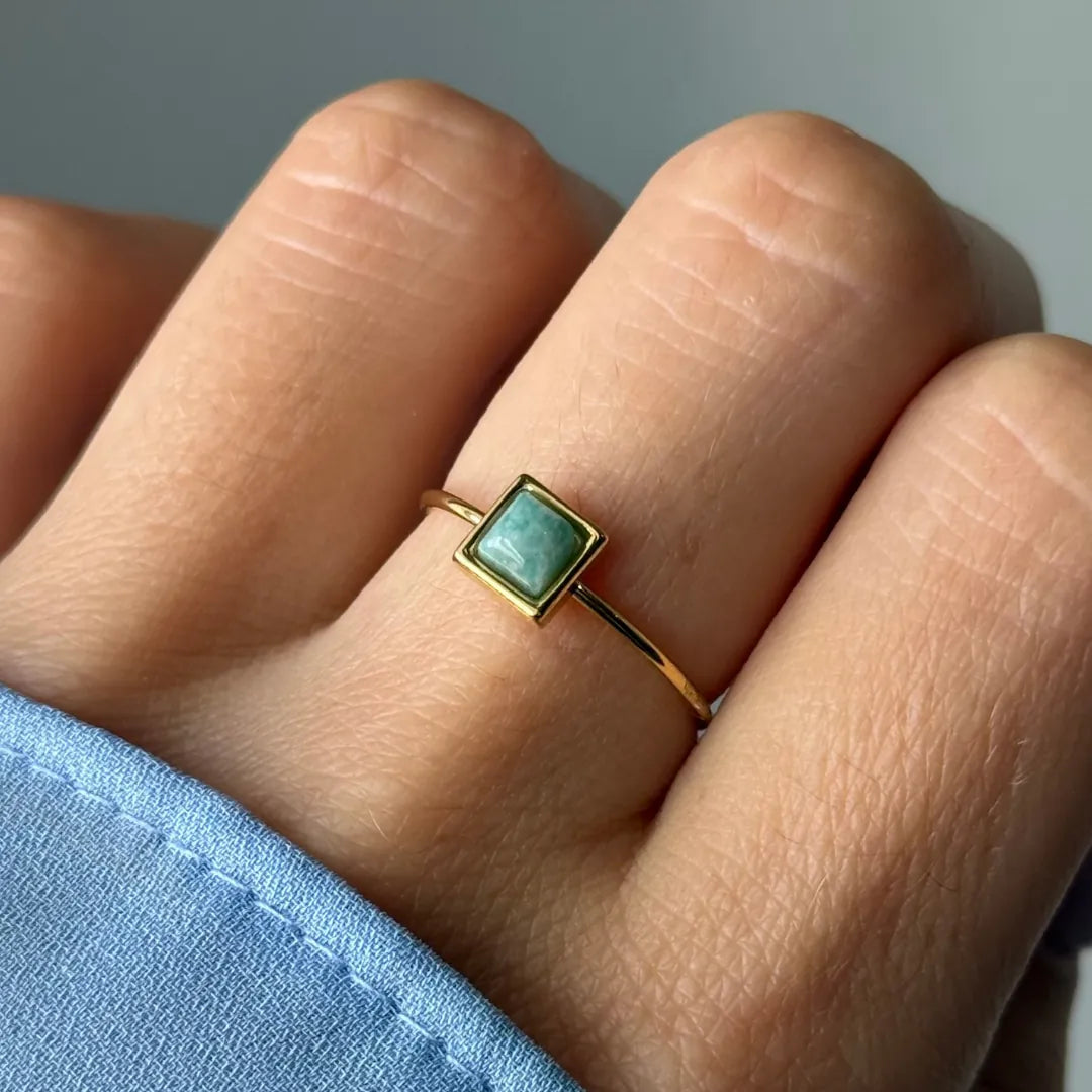Bague fine dorée ornée d'une pierre vert-bleu clair en forme de losange, portée sur un doigt avec une manche bleue.
