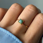 Bague fine dorée ornée d'une pierre vert-bleu clair en forme de losange, portée sur un doigt avec une manche bleue.