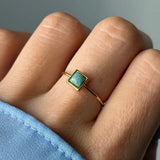 Bague fine dorée ornée d'une pierre vert-bleu clair en forme de losange, portée sur un doigt avec une manche bleue.