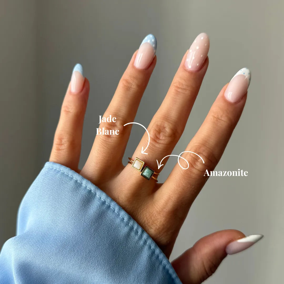Deux bagues fines dorées à pierres carrées (jade blanc, amazonite) sur un doigt d'une main à manucure bleu clair.