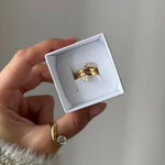 Main tenant une boîte, présentant une bague double rang en acier inoxydable doré, ornée de deux pierres de zirconium.