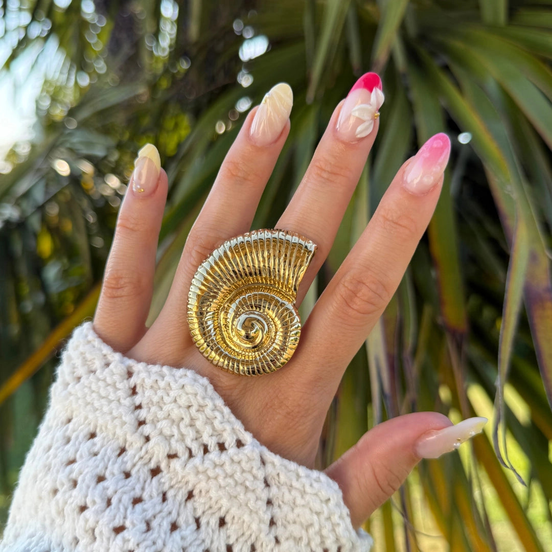 Bague acier inoxydable doré motif nautilus spiralé, portée sur une main avec ongles décorés.