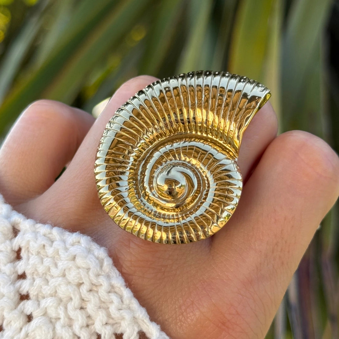 Bague en acier inoxydable doré, motif coquillage ammonite spiralé détaillé, portée sur un doigt.