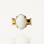 Bague dorée sertie d'une large pierre blanche ovale aspect givré ou craquelé avec fixation à griffes sur fond blanc.