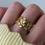 Bague en acier inoxydable doré avec un motif floral de petites fleurs, portée au doigt sur un pull jaune.