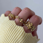Bague florale en acier inoxydable doré, motif fleurs avec petites pierres, portée sur main avec pull jaune clair.