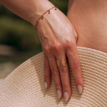 Bague ouverte en acier inoxydable doré motif coquillage sur main et chapeau de paille.