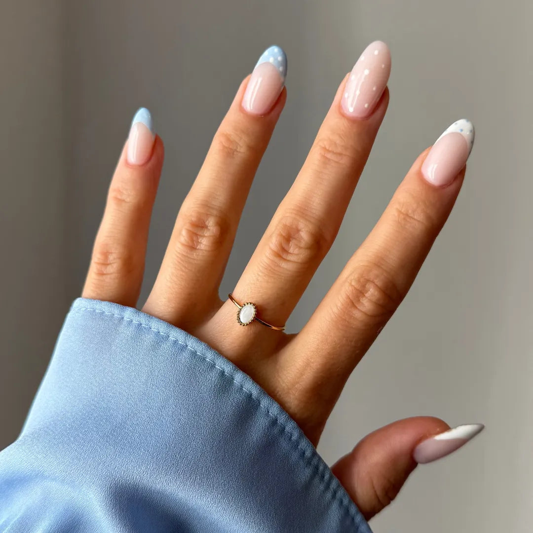 Main portant une bague fine dorée avec nacre ovale. Ongles longs style French manucure à pois et bouts colorés sur manche