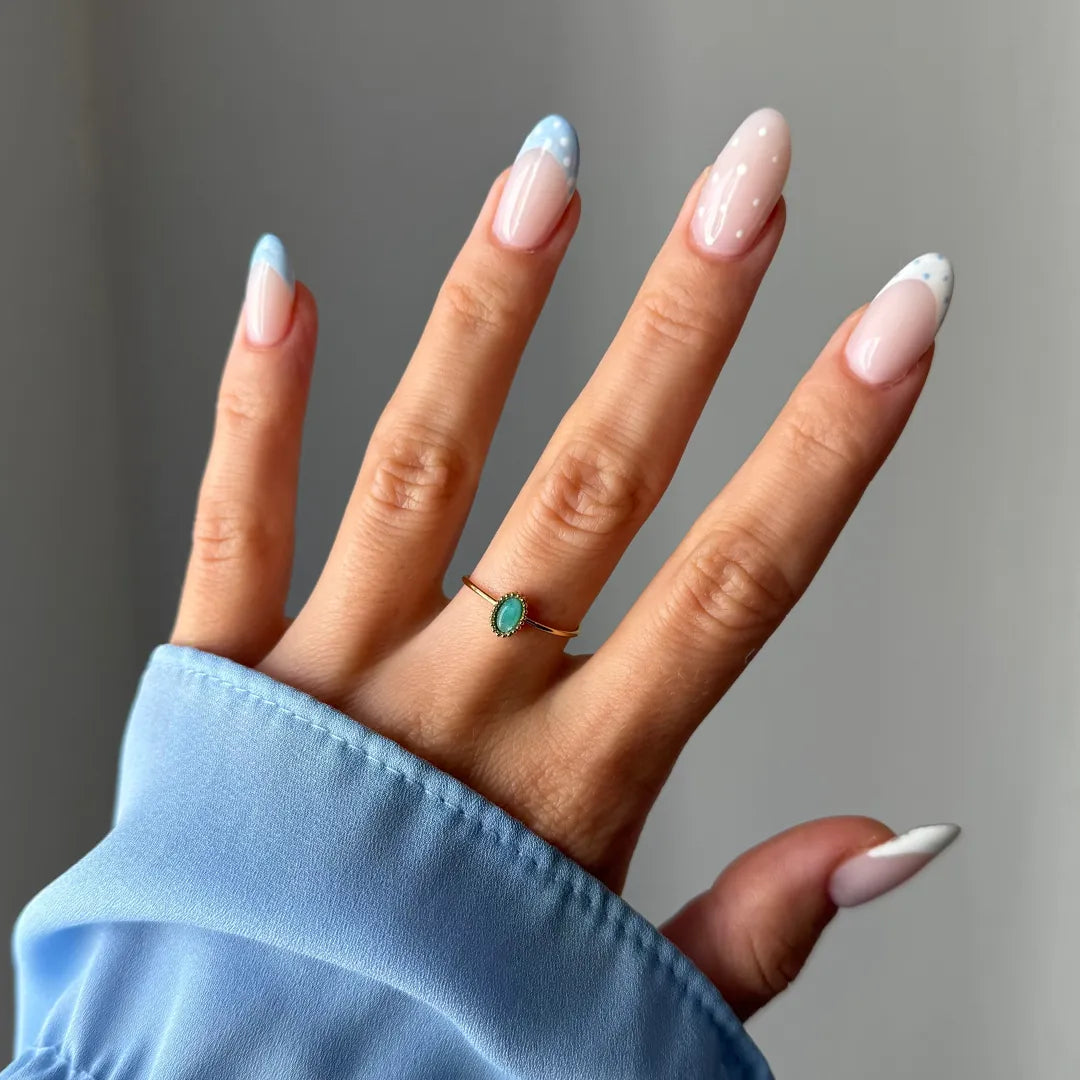 Main d'une femme portant une bague fine dorée à pierre ovale turquoise. Manchette bleue et ongles à manucure française bleu