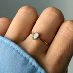 Main portant une bague fine dorée avec une pierre ovale blanche nacrée sertie de petits grains, sur une manche bleue.