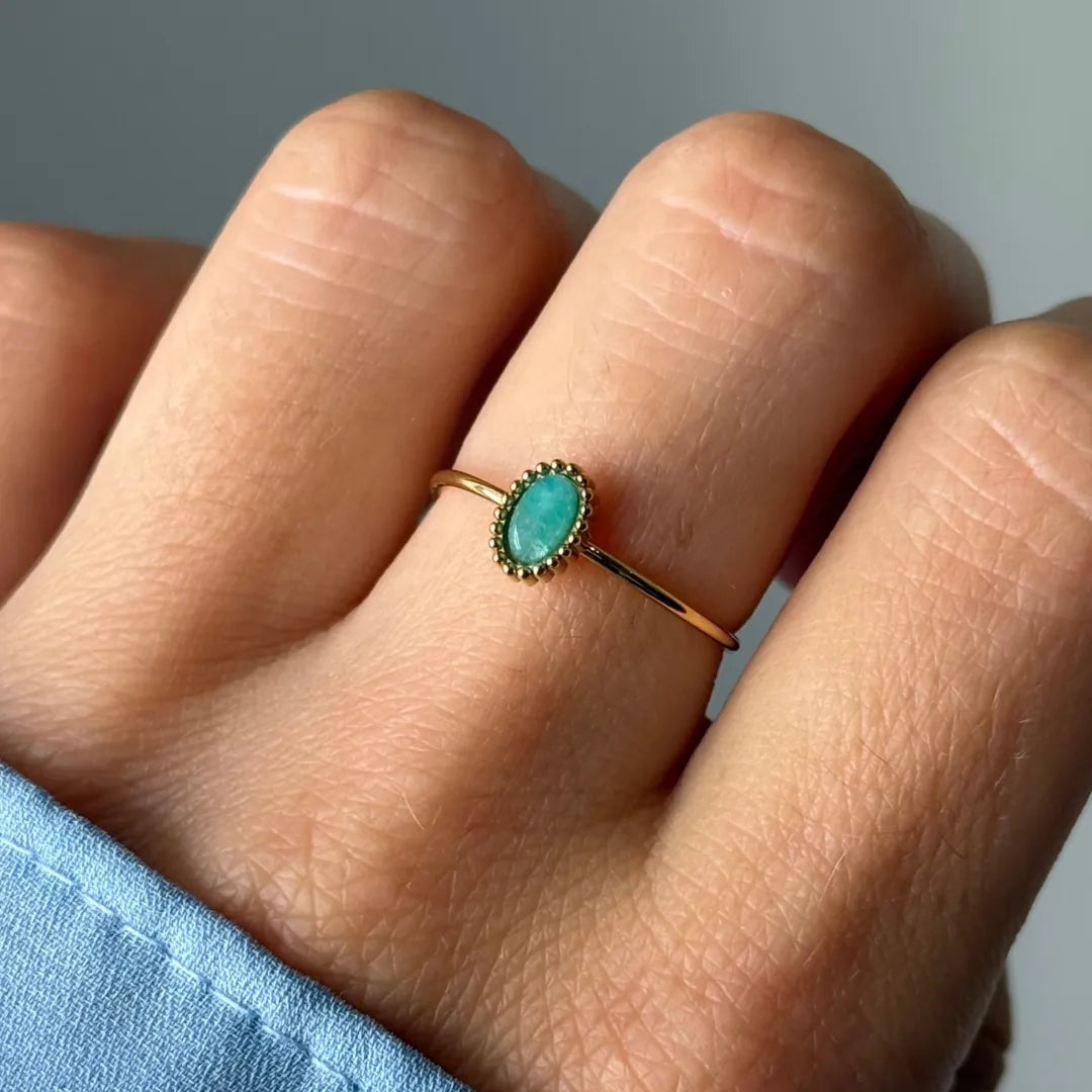 Bague fine dorée sur un doigt, avec une pierre ovale vert-turquoise encadrée d'une bordure perlée dorée.