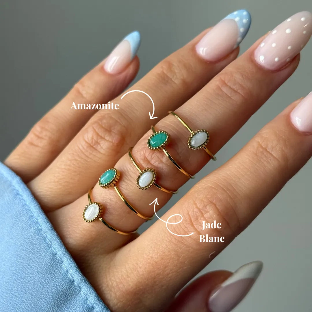 Main portant cinq bagues fines dorées avec des pierres ovales : amazonite vert-bleue et jade blanc. Ongles en manucure