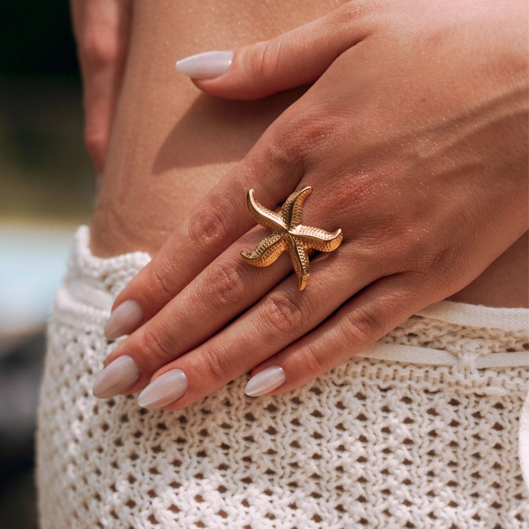 Bague en acier inoxydable dorée, motif étoile de mer, portée par une main de femme sur un vêtement blanc à mailles crochet.