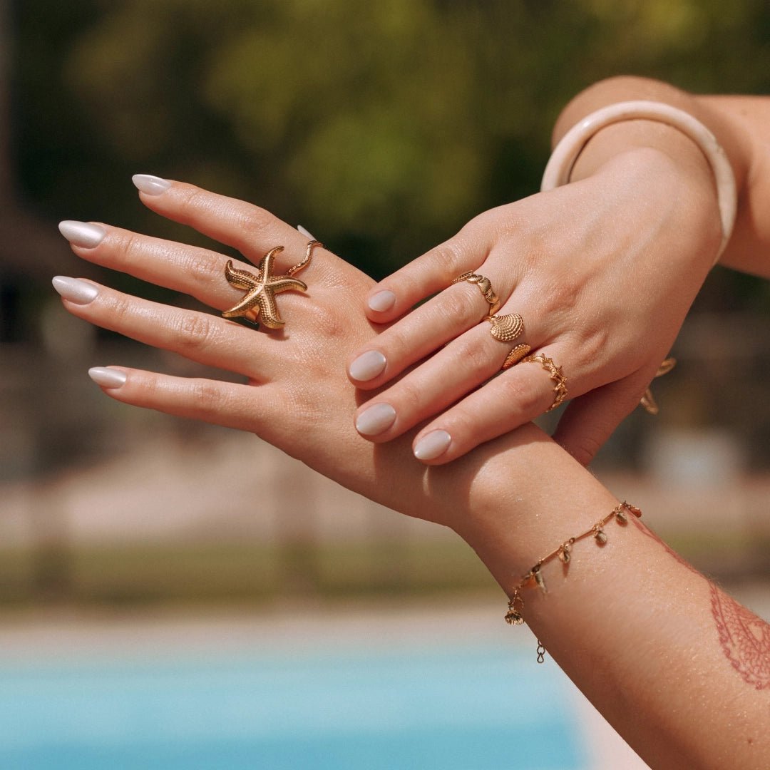 Bagues en acier inoxydable doré: étoile de mer, coquillage, petites étoiles. Mains féminines, bord de piscine en été.