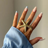 Main aux ongles bleus et blancs portant une grande bague étoile de mer texturée en acier inoxydable doré et une petite bague