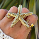 Bague en acier inoxydable dorée, grande étoile de mer texturée, portée avec d'autres bagues étoiles de mer.