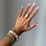 Main manucurée portant une bague en acier inoxydable doré, lisse et ondulée, une bague en nacre et des bracelets en perles.