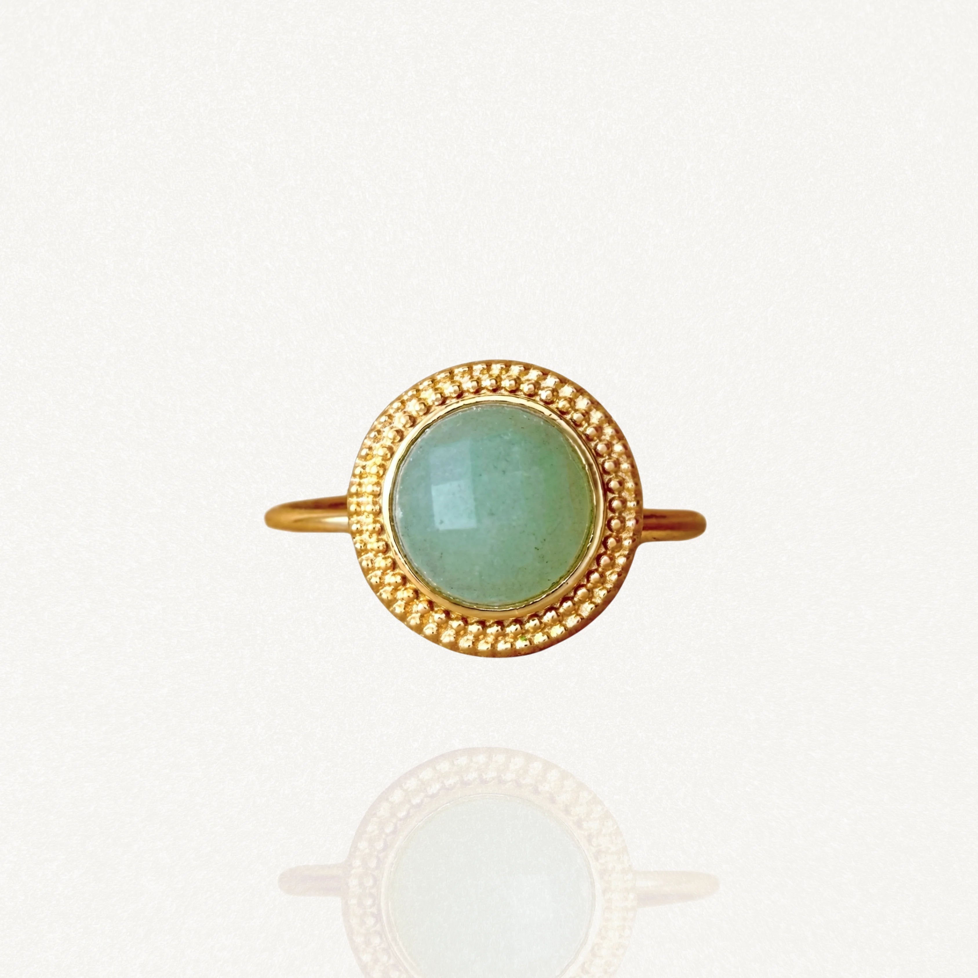 Bague fine dorée sertie d'une pierre verte ronde facettée avec un contour effet perlé, sur fond blanc.
