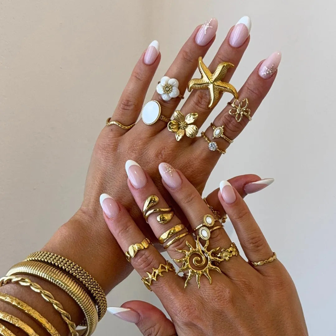 Mains aux ongles manucurés, portant de multiples bagues et bracelets en acier inoxydable doré, dont une fleur de cerisier et