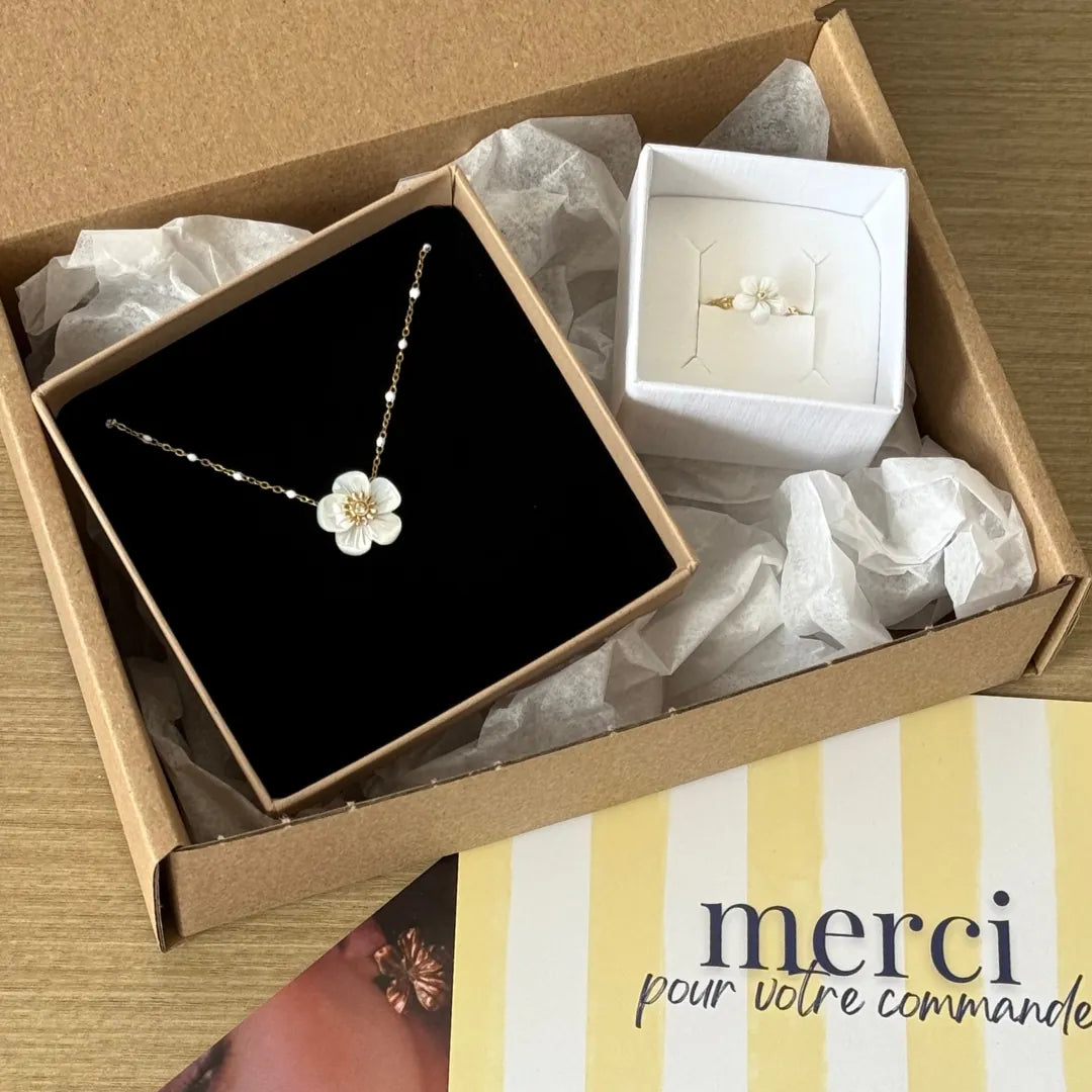 Dans une boîte cartonnée, une bague en acier inoxydable dore ornée d'une fleur en nacre, et un collier à perles assorti.