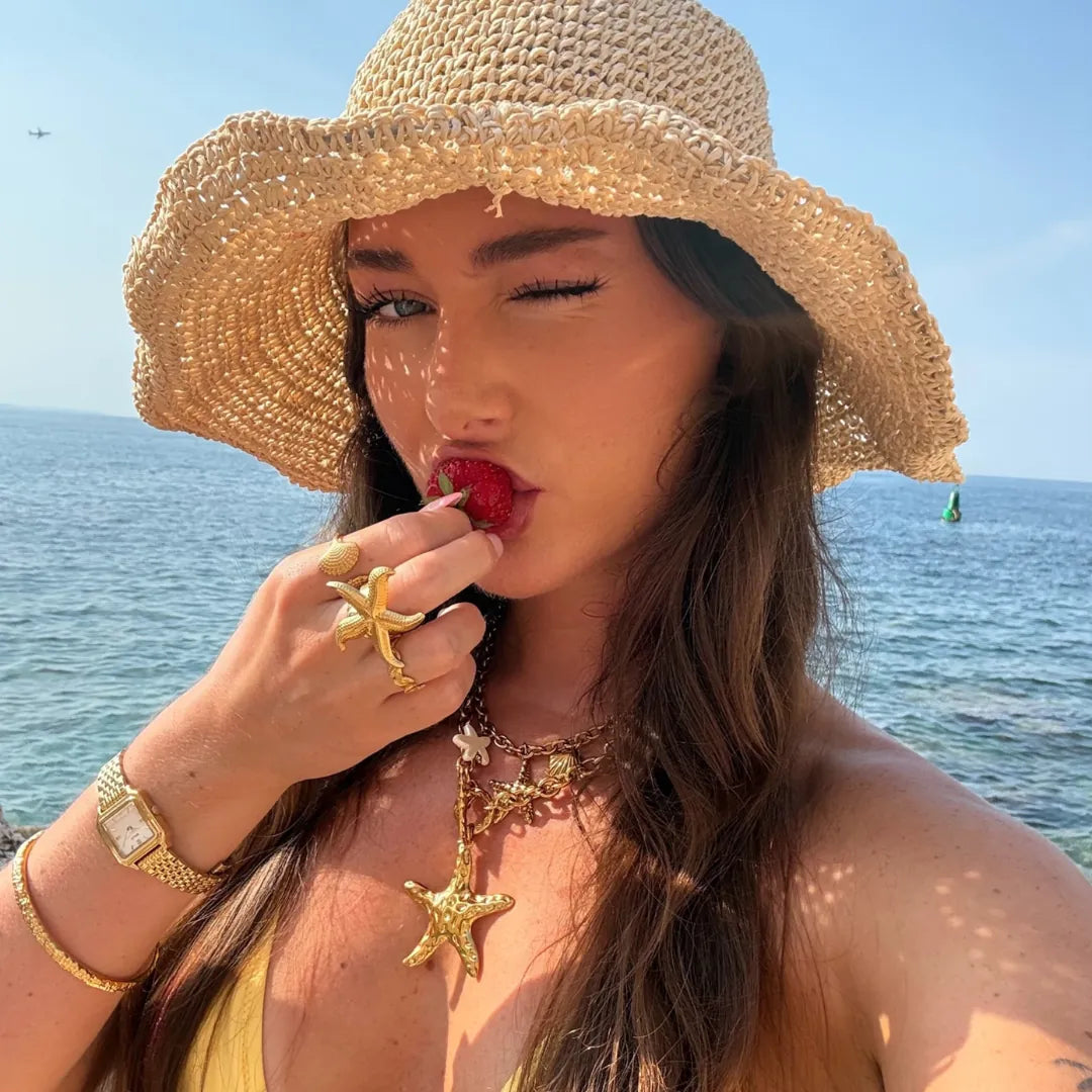 Bague en acier inoxydable doré motif étoile de mer sur la main d'une femme tenant une fraise, face à la mer.