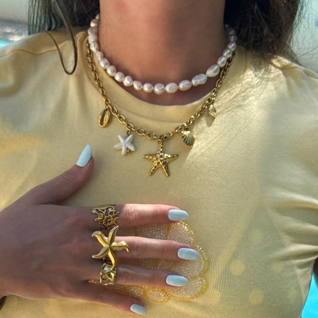 Main d'une femme avec bague étoile de mer en acier inoxydable doré, colliers de perles et breloques marines dont une étoile
