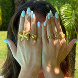 Bague large en acier inoxydable doré, avec une étoile de mer et des motifs de corail, portée par une personne aux ongles