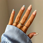 Main féminine au chemisier bleu clair, parée de trois bagues en acier inoxydable doré avec des ornements. Une bague est