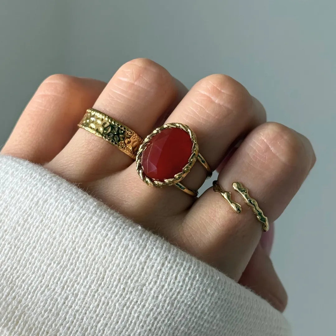 Bague en acier inoxydable doré avec une pierre naturelle ovale facettée rouge entourée d'une bordure tressée.