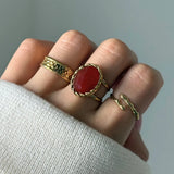 Bague en acier inoxydable doré avec une pierre naturelle ovale facettée rouge entourée d'une bordure tressée.