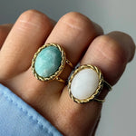 Deux bagues dorées sur un doigt, avec des pierres ovales. L'une turquoise texturée, l'autre blanche facettée, toutes deux