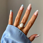 Main avec bague dorée à pierre ovale blanche facettée et bordure tressée. Ongles manucurés, certains à pointe bleue à pois