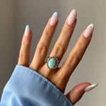 Main montrant une bague dorée à pierre ovale bleu-vert entourée d'une torsade. Manche de chemise bleue et ongles avec motifs