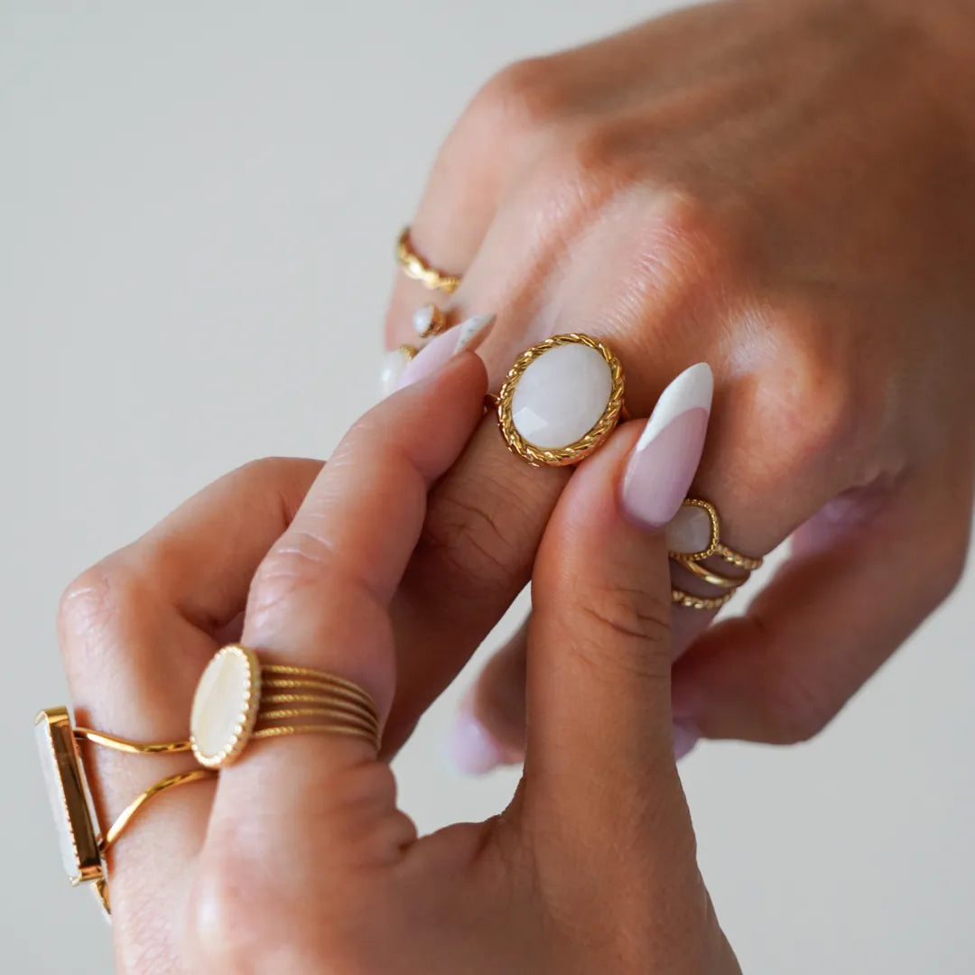 Mains portant plusieurs bagues dorées. L'une est ovale avec une pierre blanche laiteuse et un bord torsadé. Ongles manucurés.