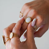 Mains portant plusieurs bagues dorées. L'une est ovale avec une pierre blanche laiteuse et un bord torsadé. Ongles manucurés.