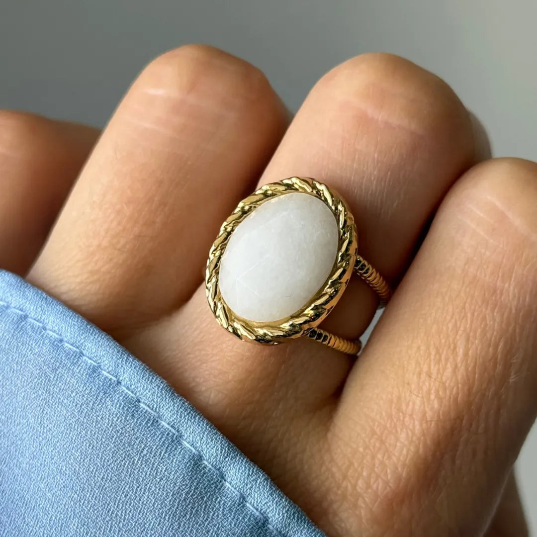 Bague dorée portée au doigt, avec une pierre ovale blanche légèrement veinée sertie d'un cadre tressé, et un anneau double