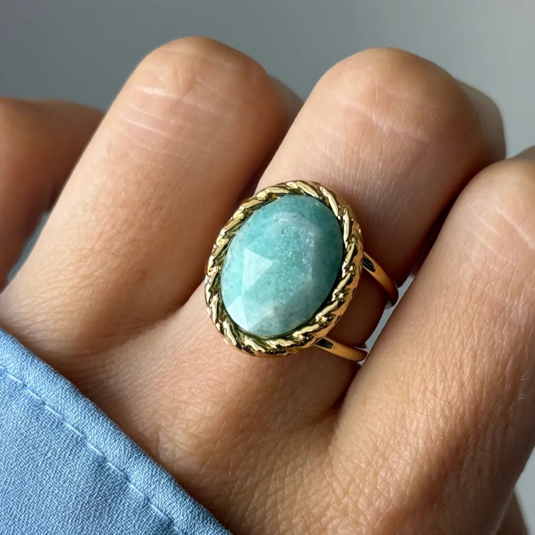 Bague dorée sur un doigt, avec une pierre ovale bleu-vert clair facettée entourée d'un motif tressé. Manche bleue.