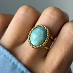 Bague dorée sur un doigt, avec une pierre ovale bleu-vert clair facettée entourée d'un motif tressé. Manche bleue.