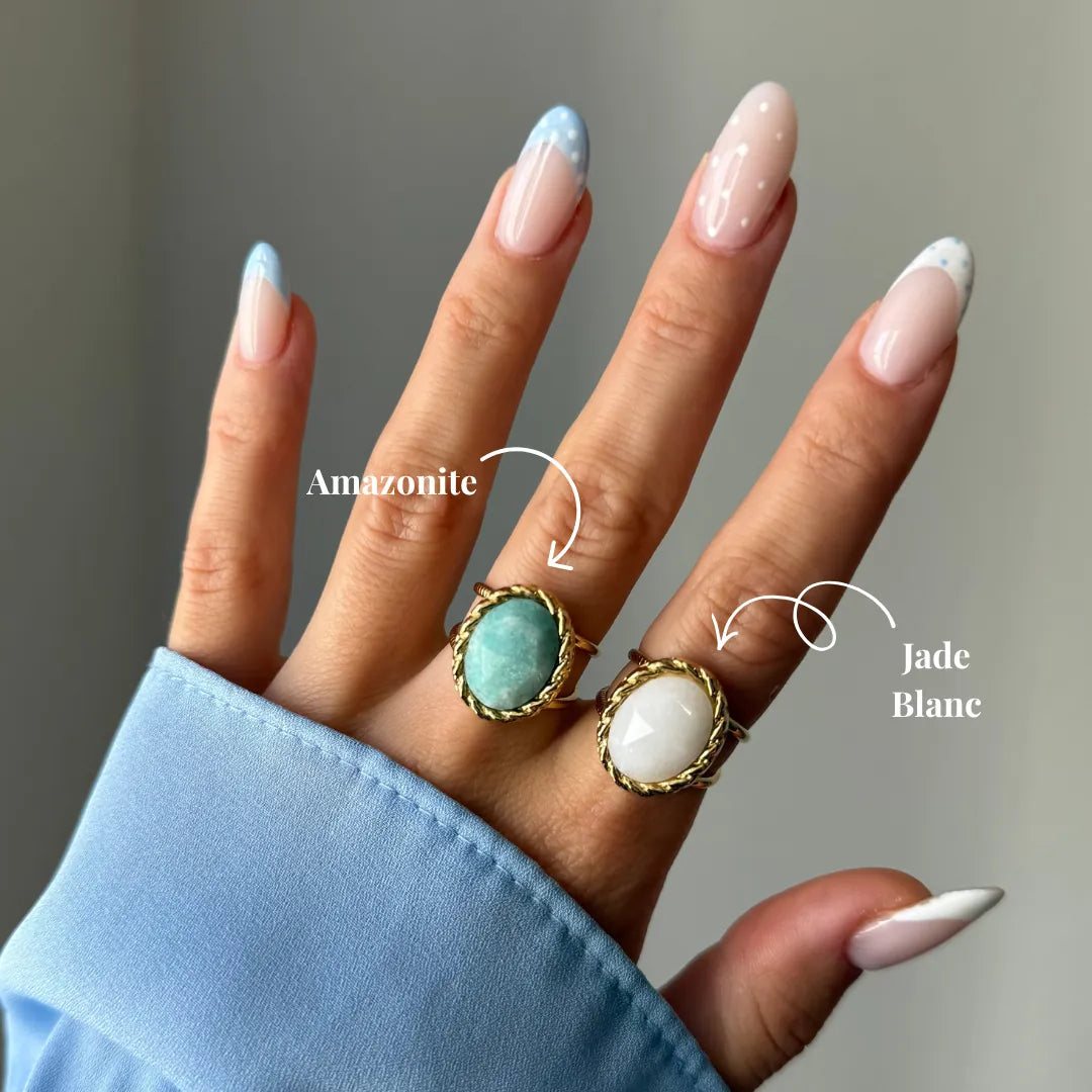 Main avec deux bagues à pierre ovale: amazonite turquoise et jade blanc. Montures dorées torsadées. Ongles manucurés bleu et