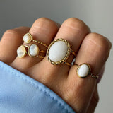 Cinq bagues dorées avec cabochons blancs sur une main. La plus grande est ovale bordée d'un tressage, d'autres sont ovales ou