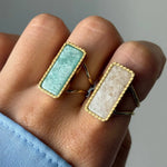 Deux bagues dorées à pierres rectangulaires, une bleu-vert et une blanc cassé, portées sur une main. Bordure perlée.