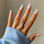 Main sur manche de chemise bleue, portant une bague en or avec pierre rectangulaire blanche laiteuse, bord perlé. Ongles