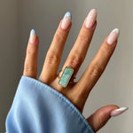 Main portant une bague dorée à pierre rectangulaire vert d'eau, sertie d'un bord perlé. Manucure française bleu ciel et