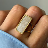 Bague dorée avec pierre rectangulaire blanche marbrée et cadre perlé portée sur une main, manche de chemise bleue visible.