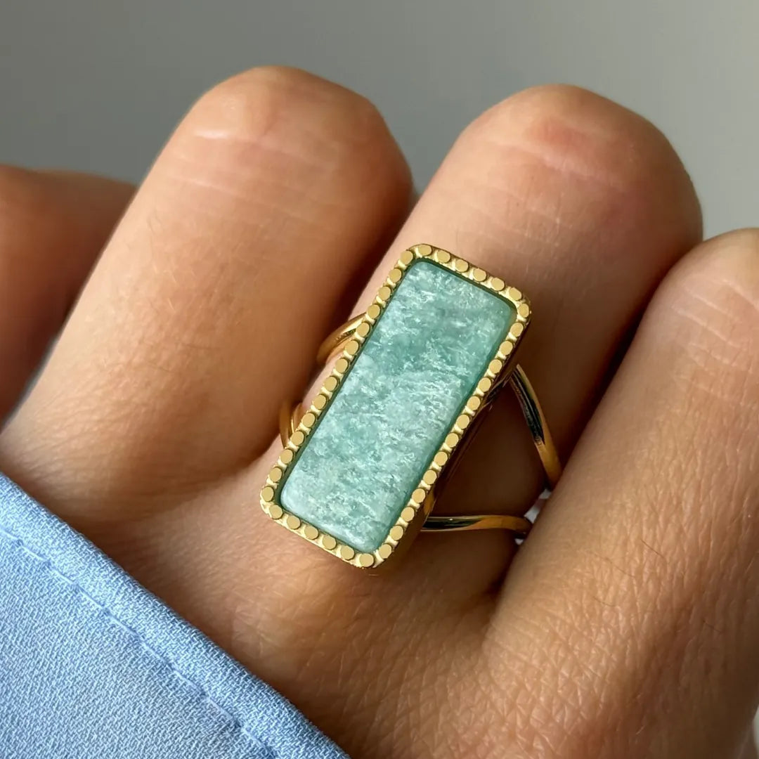 Bague dorée à pierre rectangulaire vert d'eau, encadrée d'un liseré de billes, portée sur un doigt.