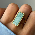 Bague dorée à pierre rectangulaire vert d'eau, encadrée d'un liseré de billes, portée sur un doigt.