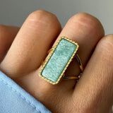 Bague dorée à pierre rectangulaire vert d'eau, encadrée d'un liseré de billes, portée sur un doigt.